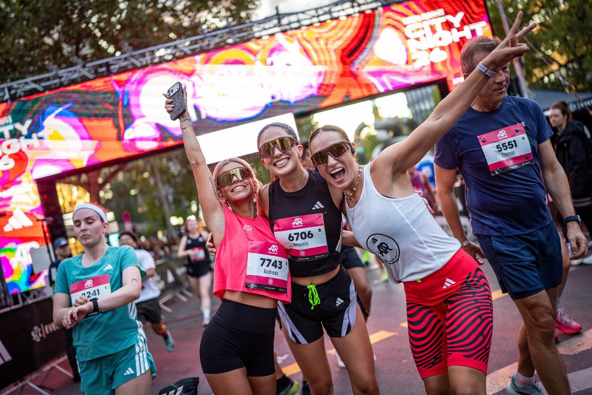 Three laughing and cheering female runners standing arm in arm at the finish line, with the illuminated finish gantry just behind them.