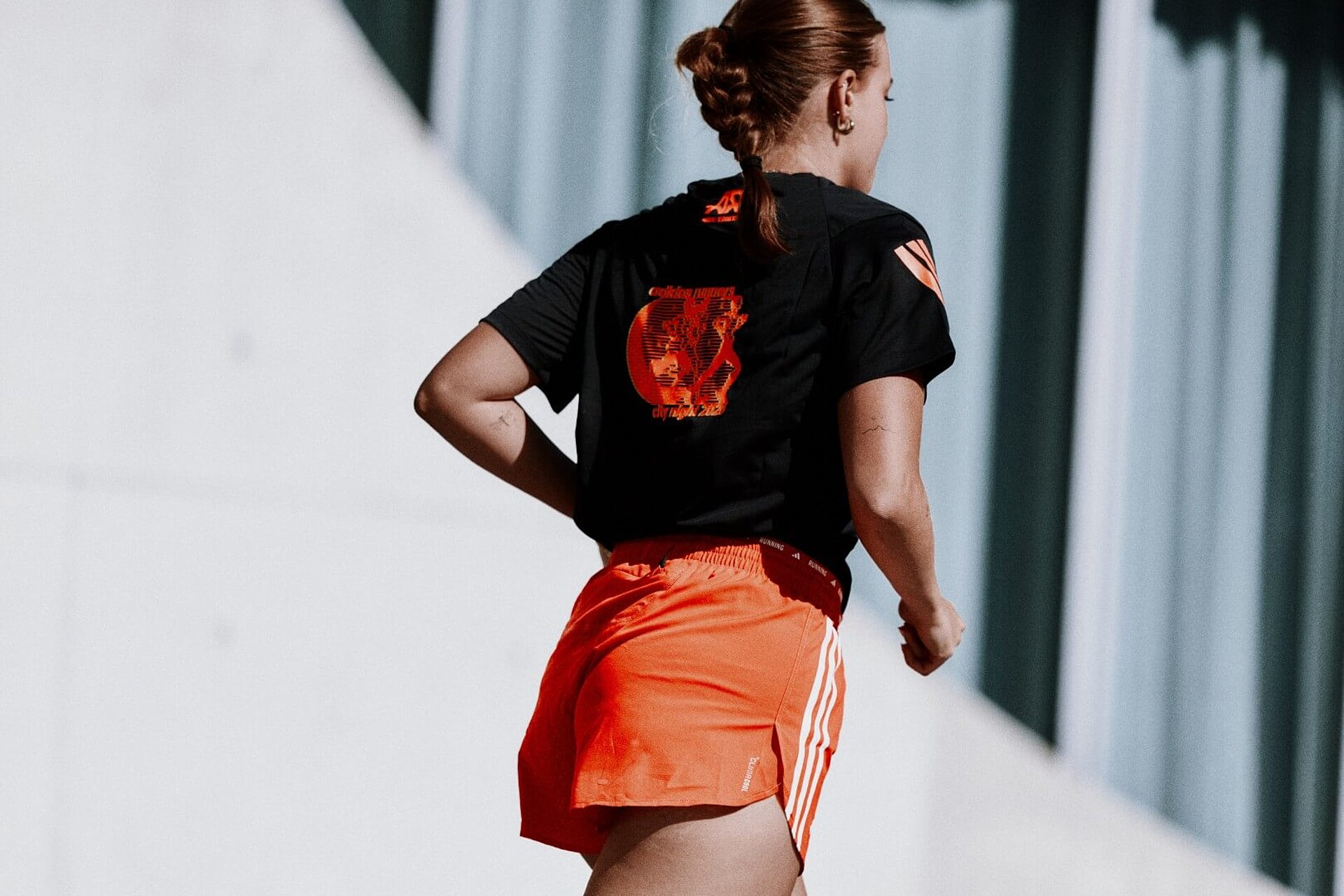 A female runner, seen from behind, wearing a black T-shirt with orange accents and orange shorts, both by adidas.